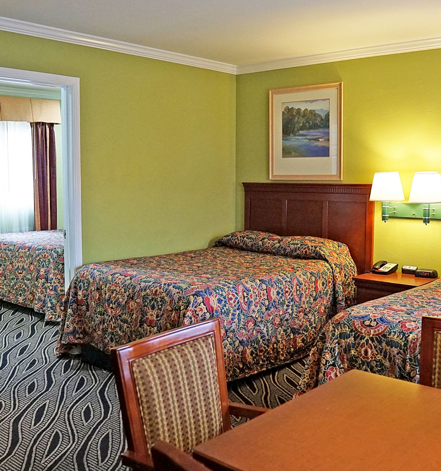 This image shows a hotel room with two beds, a table with chairs, a lamp, and a view into a bathroom with towels hanging.