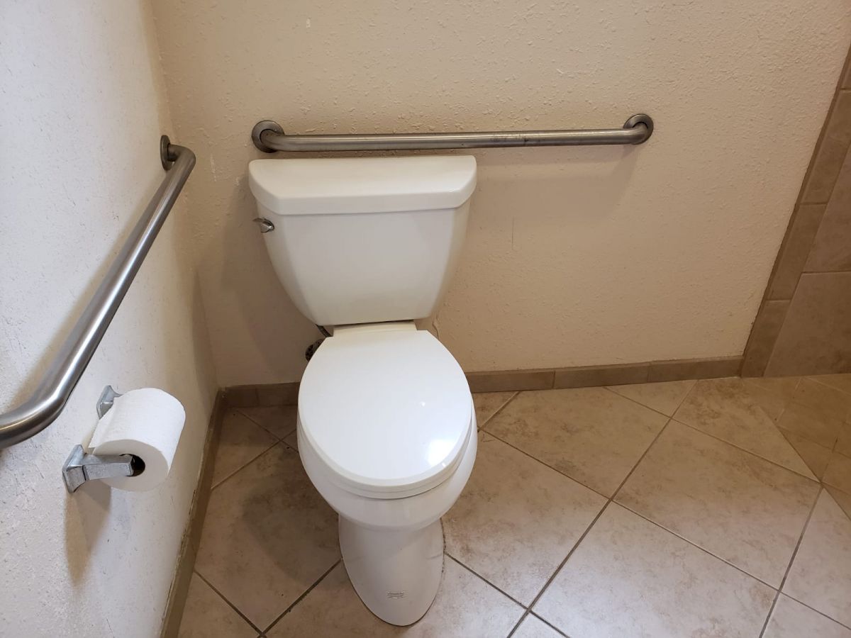 This image shows a white toilet with handrails on both sides along with a wall-mounted toilet paper holder, set against beige tiled flooring.