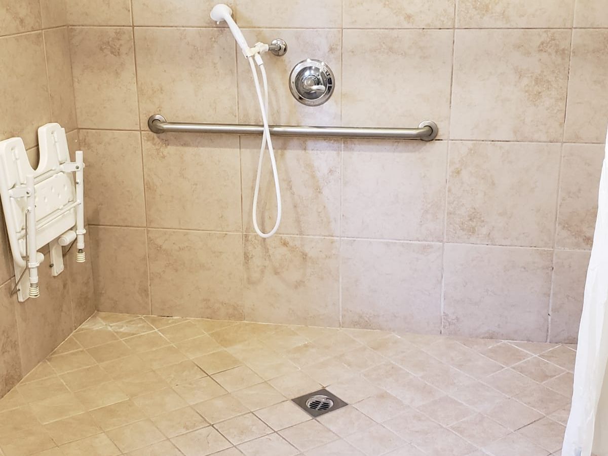 A tiled walk-in shower with a handheld showerhead, grab bar, wall-mounted folding seat, and floor drain.