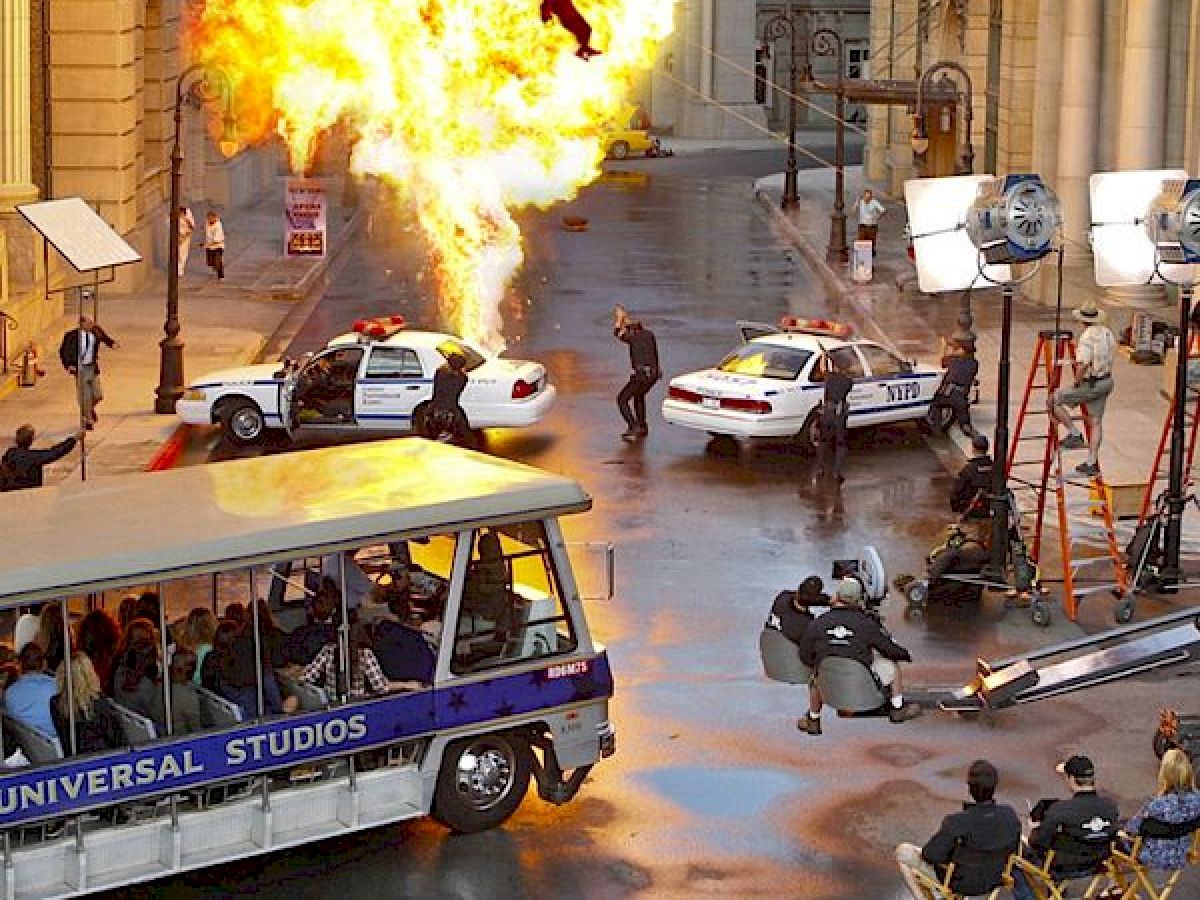 A film set with a fiery explosion scene being shot, police cars, crew, actors, and a Universal Studios tour tram with passengers observing.