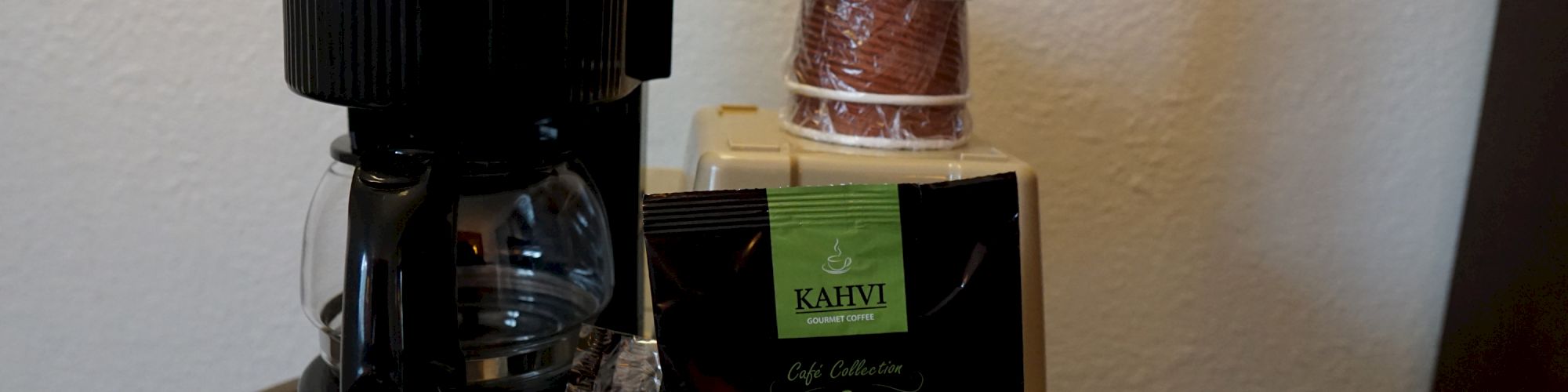 The image shows a coffee setup with a drip coffee maker, a stack of disposable cups, and a packet of Kahvi coffee on a tray.