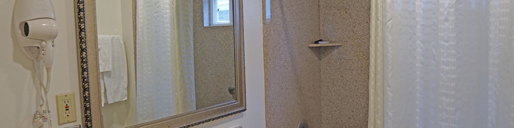 A bathroom with a sink, large mirror, wall-mounted hair dryer, and a shower with a curtain. The sink countertop has a soap dispenser.