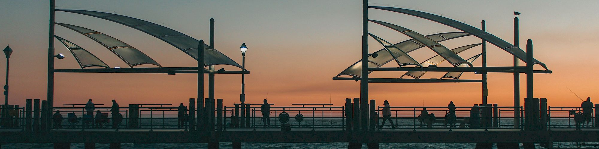 A pier extends into the water at sunset, with silhouetted people and modern canopy structures, against a colorful sky and illuminated lamp posts.