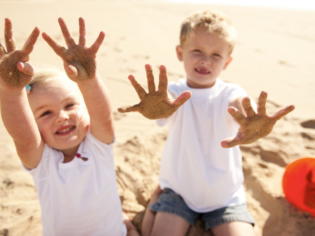 Two children are sitting on the beach, showing their sandy hands and smiling. Nearby, there is an orange bucket partially buried in the sand.