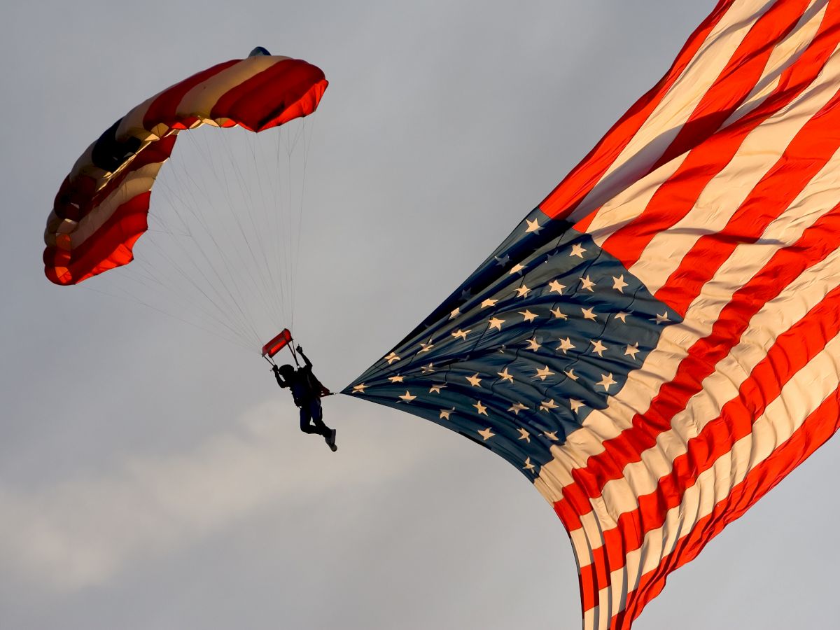 A person is parachuting, holding onto a large American flag that is trailing behind them in the sky.