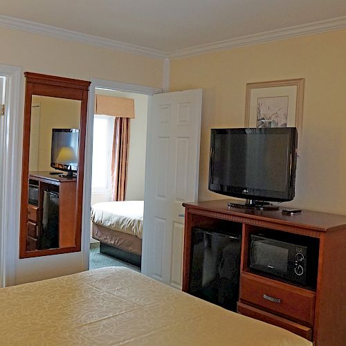 A hotel room with a bed, a wooden dresser holding a TV and microwave, a bathroom entrance, and a view into a potentially connected room.