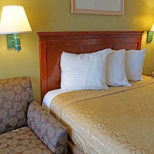 A cozy hotel room with a patterned armchair, a neatly made bed with three pillows, wall-mounted lamps, and a nightstand with a phone.