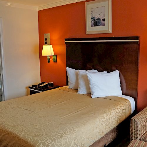 This image shows a hotel room with a bed, two lamps, an armchair, and a small bathroom with a shower visible through the door.