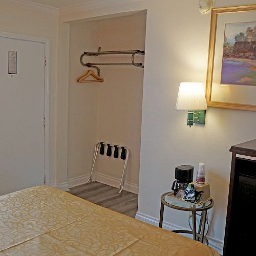 A hotel room with a bed, TV, mini-fridge, table with coffee maker, hanging rack with hangers, artwork on the wall, door, and window.