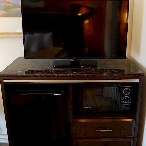 A TV on a cabinet with a microwave, mini fridge, and three drawers underneath. A framed picture is partially visible on the wall behind.