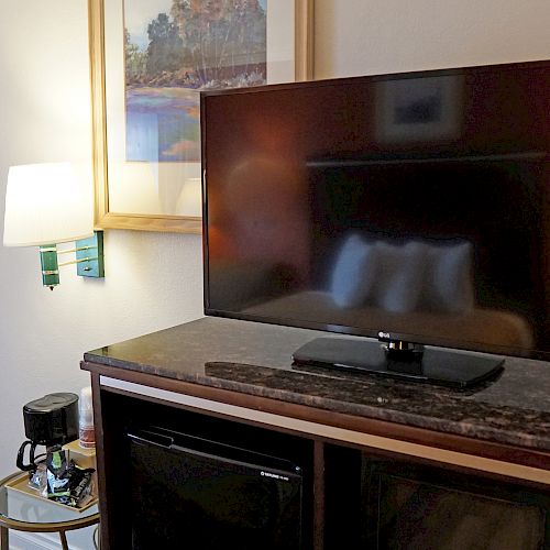 A hotel room with a mounted TV, a lamp, a painting, a small table holding a coffee maker, and an open closet with hangers is shown.