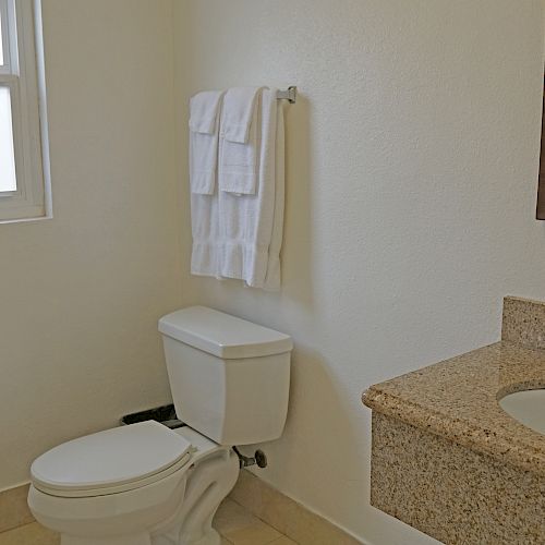 The image shows a bathroom with a toilet, a wall-mounted towel rack with folded white towels, a sink with a countertop, and a mirror above it.