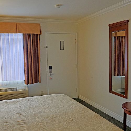 This image shows a hotel room with a bed, TV, window, lamps, air conditioner, and mirror. The decor is simple with a neutral color scheme and minimal furniture.