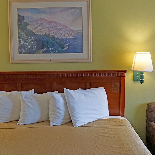 A neatly made bed with four pillows, flanked by wall-mounted lamps, a painting, and an armchair beside it within a cozy room.