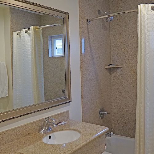 This image shows a bathroom with a sink, large mirror, hairdryer, and a bathtub with a shower curtain and window, creating a clean and neutral look.