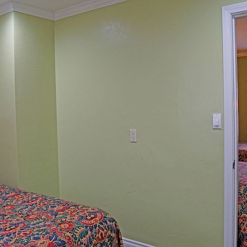 The image shows a hotel room with two beds, colorful bedspreads, light green walls, a picture frame on the wall, and an adjoining room with another bed.