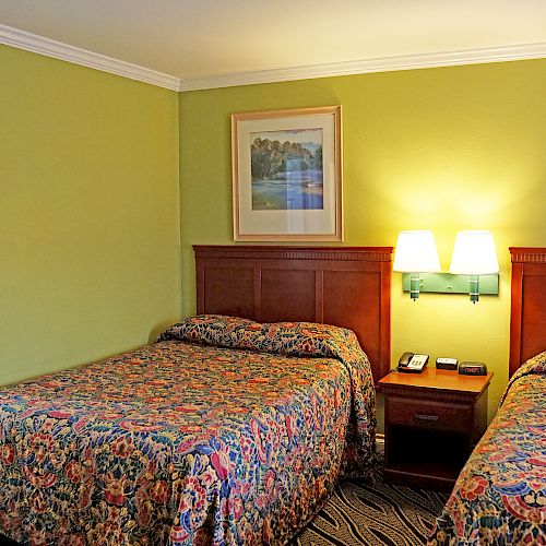 The image shows a hotel room with two double beds, a nightstand with a phone, two framed pictures above the beds, and a doorway to another room.