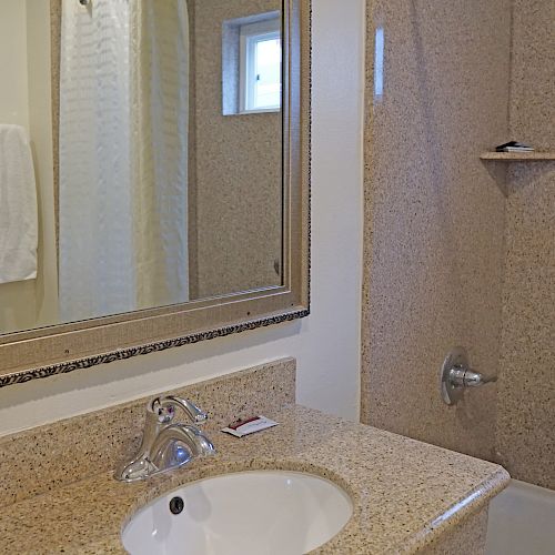 A bathroom with a granite countertop, sink, mirror, towel rack with towels, and a bathtub-shower combo with a small window and shelf.