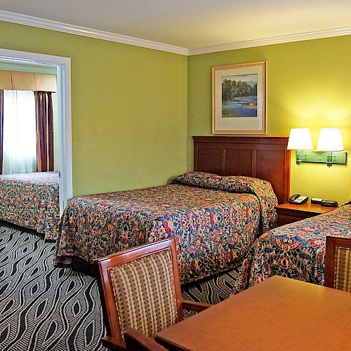 A hotel room with two beds, a lamp, a table with chairs, patterned bedspreads, framed pictures on the wall, and a doorway leading to a bathroom.