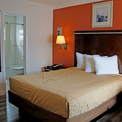 A hotel room featuring a bed with an orange wall, two lamps, a small armchair, and a bathroom visible through an open door.