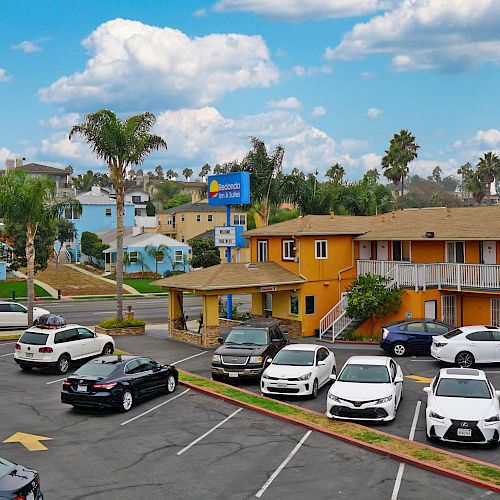 A parking lot with various cars is adjacent to a row of colorful buildings, palm trees, and a bright, cloudy sky in the background.