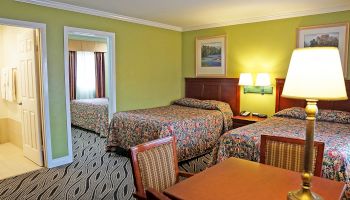 This image shows a hotel room with two beds, a table with chairs, a lamp, artwork on the walls, and access to a bathroom with towels.