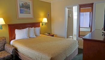 A cozy hotel room with a large bed, chair, dresser, TV, lamps, artwork, and a bathroom.