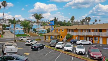 A parking lot with several cars, surrounded by colorful buildings, palm trees, and a clear sky with clouds, possibly a motel or apartment complex.