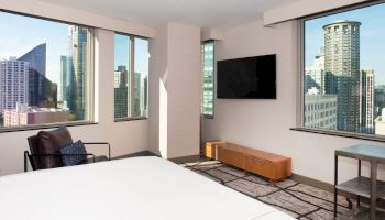 A modern hotel room with large windows showcasing a cityscape view, a wall-mounted TV, and simple furnishings.