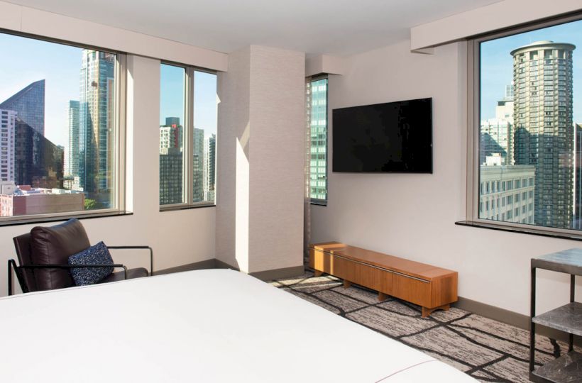 A modern hotel room with large windows showcasing a cityscape view, a wall-mounted TV, and simple furnishings.