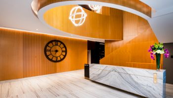A modern lobby with a marble reception desk, large wooden clock, spiral staircase, and decorative lights, accented by a vase of colorful flowers.