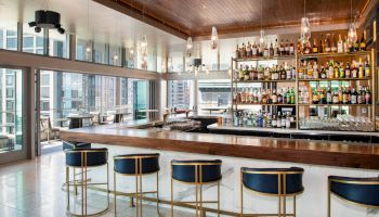 The image shows a modern bar with a sleek wooden counter, gold-accented barstools, and shelves stocked with various bottles of alcohol.