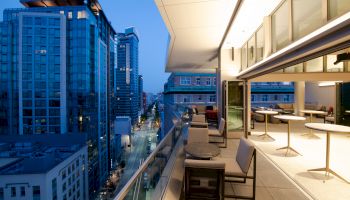 This image shows a modern cityscape with tall buildings and a spacious balcony with seating and tables overlooking a bustling street below.