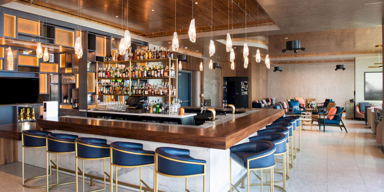 The image shows a modern bar with a wooden counter, blue chairs, various bottles on shelves, and hanging lights, set in a spacious, elegant interior.