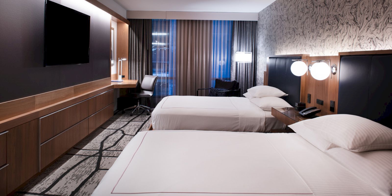 The image shows a modern hotel room with two neatly made beds, a desk with a chair, a wall-mounted TV, and floor-to-ceiling windows with curtains.