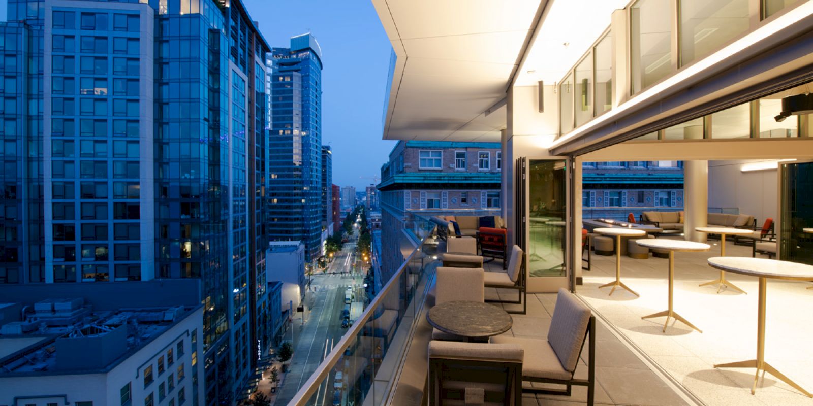 A modern balcony overlooking a city skyline at dusk, featuring outdoor seating and tables, with tall buildings illuminated by streetlights below.