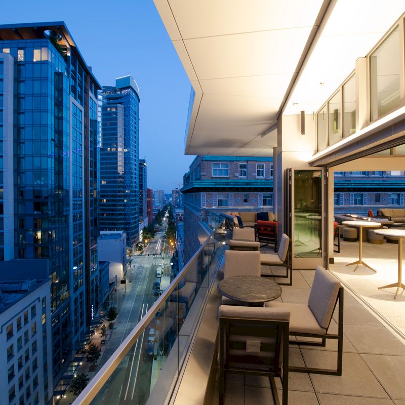A modern balcony with seating and tables overlooking a cityscape filled with tall buildings at dusk, featuring a mix of glass and concrete structures.