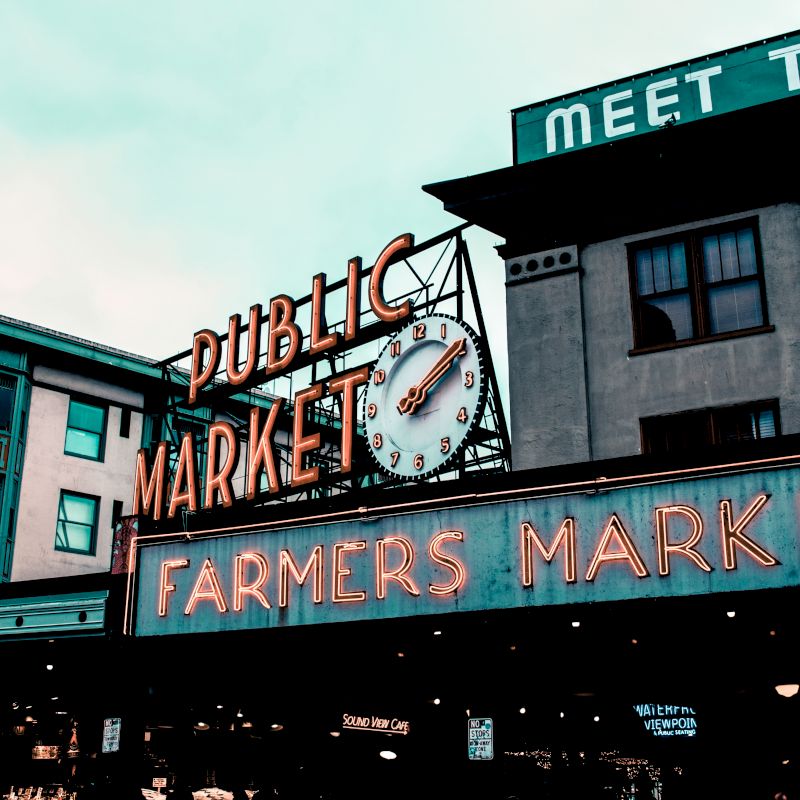 This image shows the entrance to a public farmers market with neon signs and a clock. Buildings and a hotel sign are visible in the background.