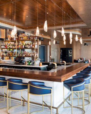 A modern bar with a wooden counter, blue chairs, hanging light fixtures, and a stocked bar shelf in a stylish, spacious interior.