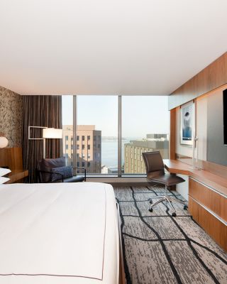 A modern hotel room features a large bed, a wall-mounted TV, a desk with a chair, and a view of buildings outside the window.