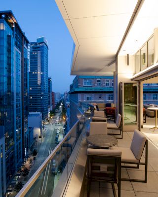 This image shows a modern cityscape at dusk with tall buildings and an elegant outdoor terrace featuring tables, chairs, and lighting.