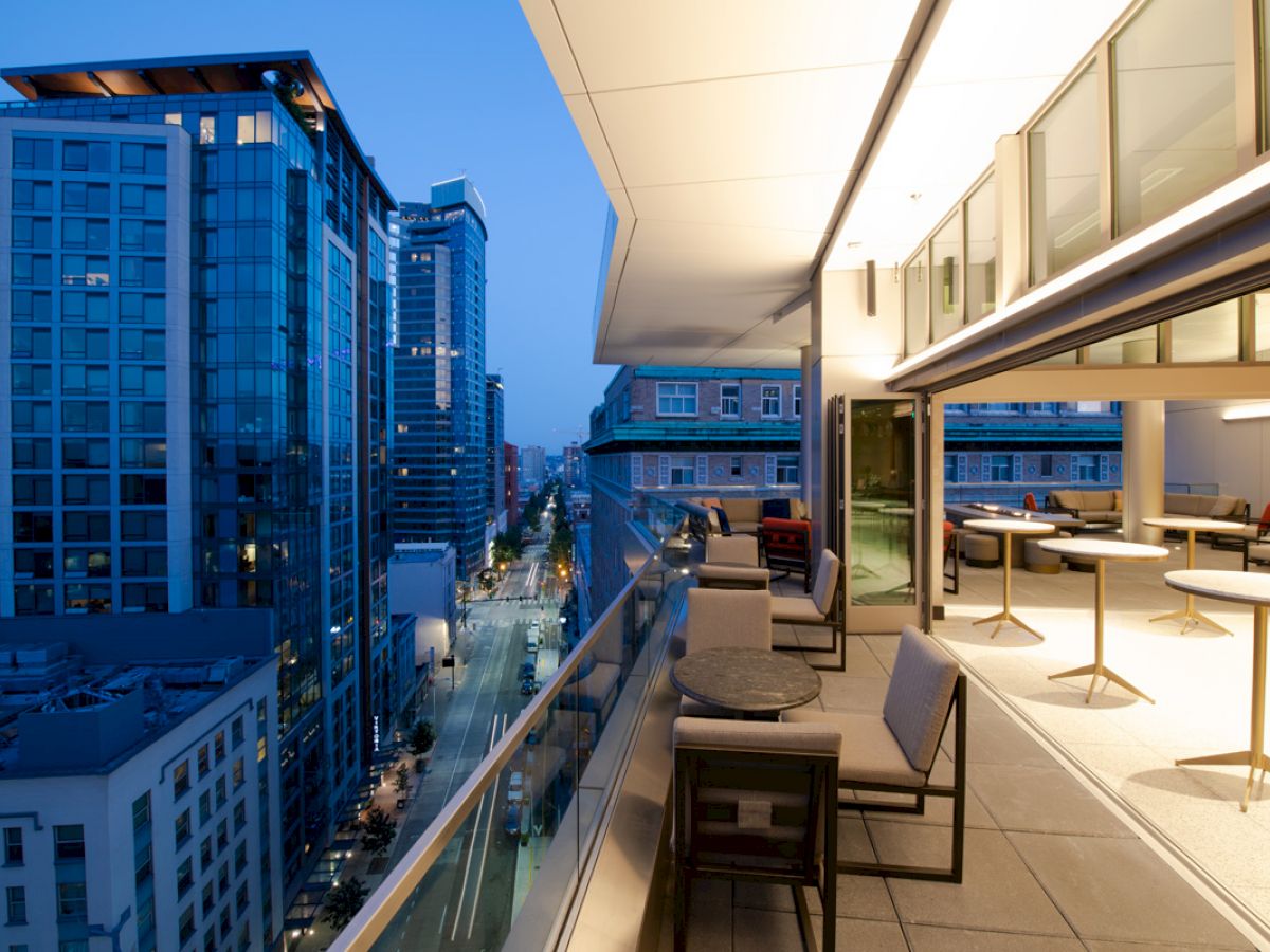 A modern balcony with seating overlooks a city street lined with high-rise buildings at dusk, featuring bright interior lighting and a sleek design.