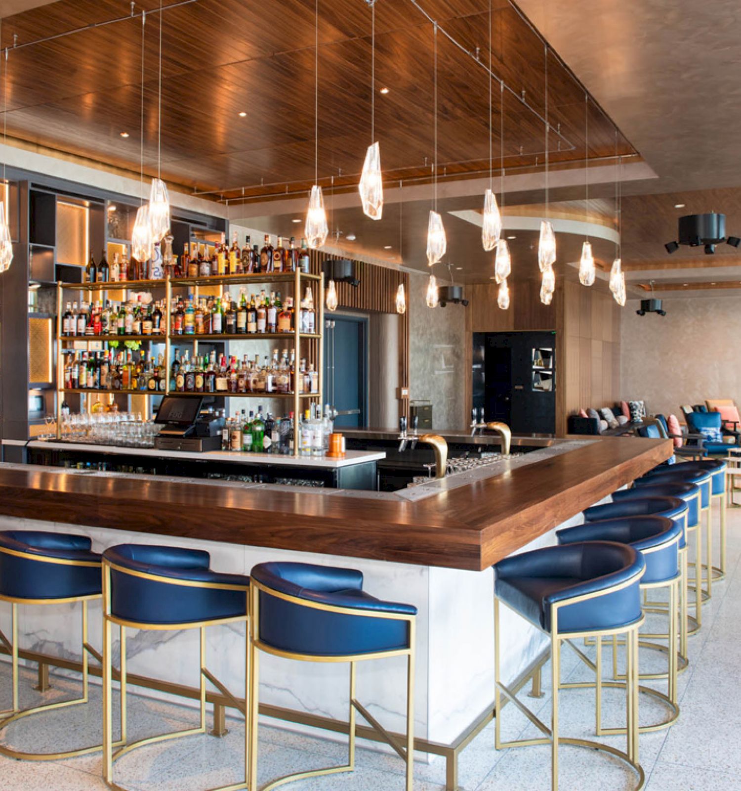 This image depicts a modern bar with blue cushioned bar stools, hanging pendant lights, and a well-stocked liquor shelves.