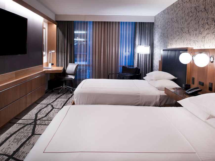 A modern hotel room with two neatly made beds, a TV, a desk with a chair, and large windows with curtains.