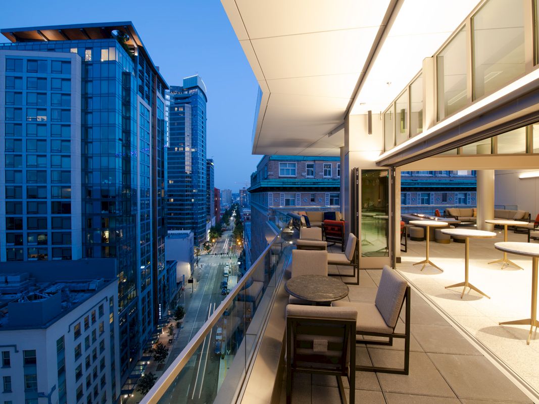 The image shows a modern cityscape viewed from a terrace during early evening, featuring high-rise buildings, street below, and outdoor seating.