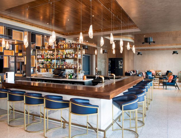 The image depicts a modern, stylish bar with a central counter, shelves stocked with various bottles, bar stools, and ambient lighting fixtures.