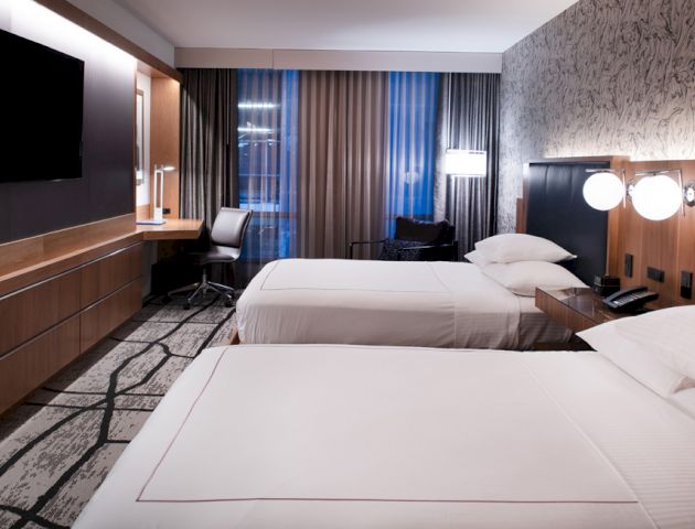 A modern hotel room with two beds, a flat-screen TV, a desk with a chair, and minimalist decor; ambient lighting and large window with curtains.
