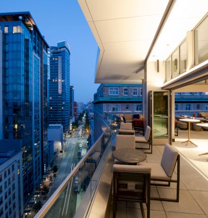 A modern city view from an elevated terrace at dusk, with tables and chairs arranged along the balcony, adjacent to a lit indoor space.