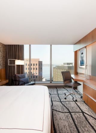 This image shows a modern hotel room with a large bed, desk, chair, TV, and a window with a city and water view in the background.