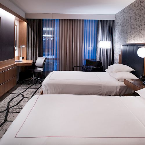 A modern hotel room with two beds, a wall-mounted TV, a desk with a chair, and stylish decor featuring a large window with curtains.